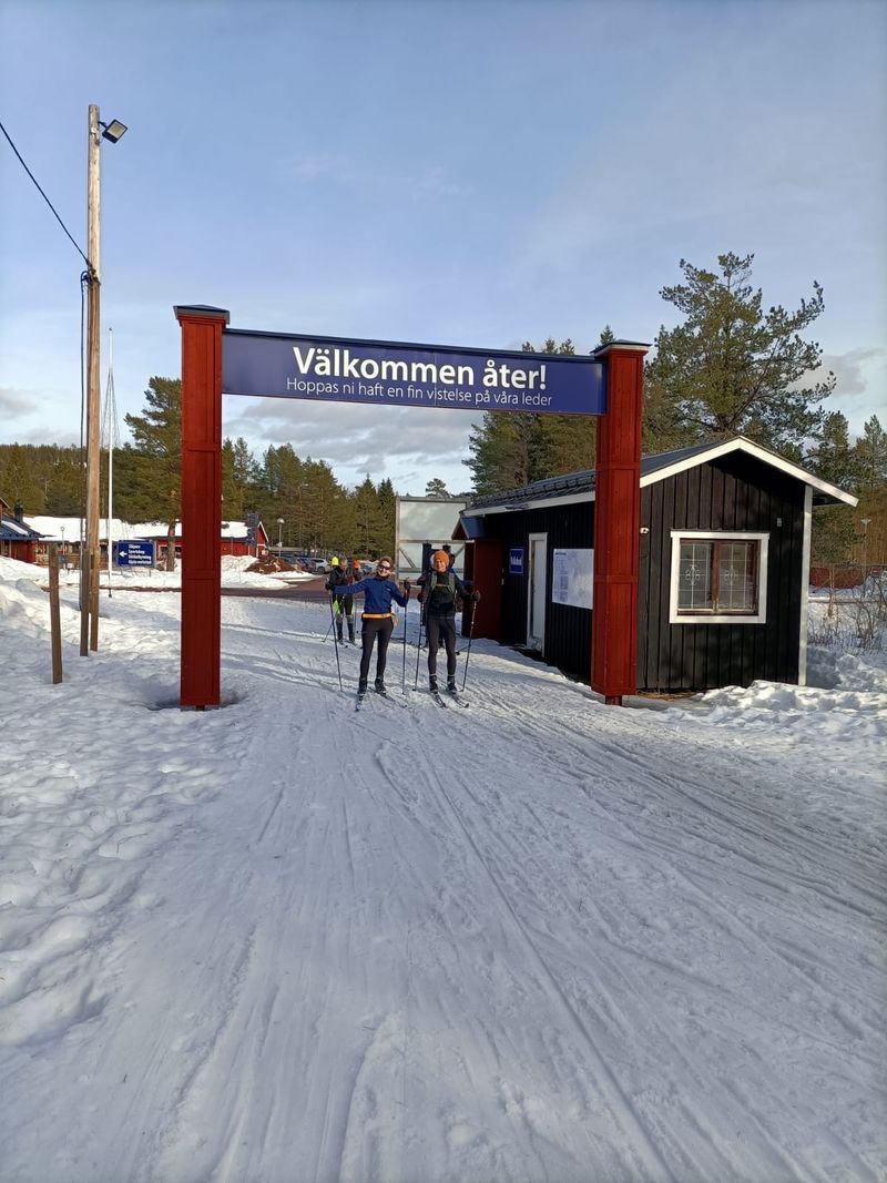 Twee langlaufers onder een rode houten poort met het bord Välkommen åter, onder een blauwe lucht