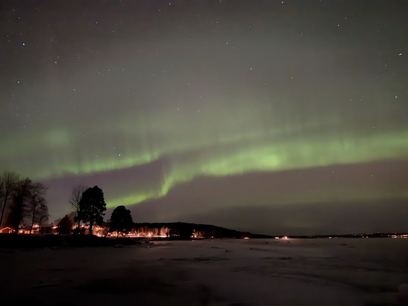 Groen noorderlicht boven een bevroren meer, met lichtjes langs de oever
