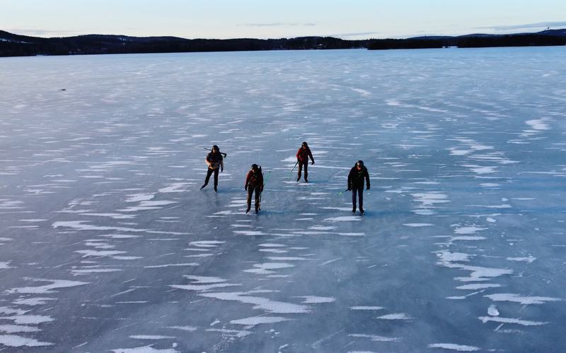 Luchtfoto van vier schaatsers op een groot bevroren meer met sneeuwstrepen op het ijs en een bosrand aan de horizon