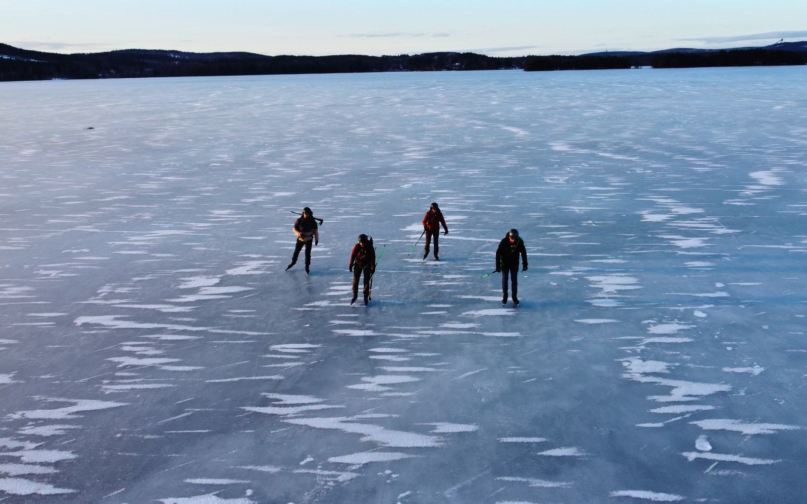 Vier schaatsers op een groot meer met zwart natuurijs, van bovenaf gezien