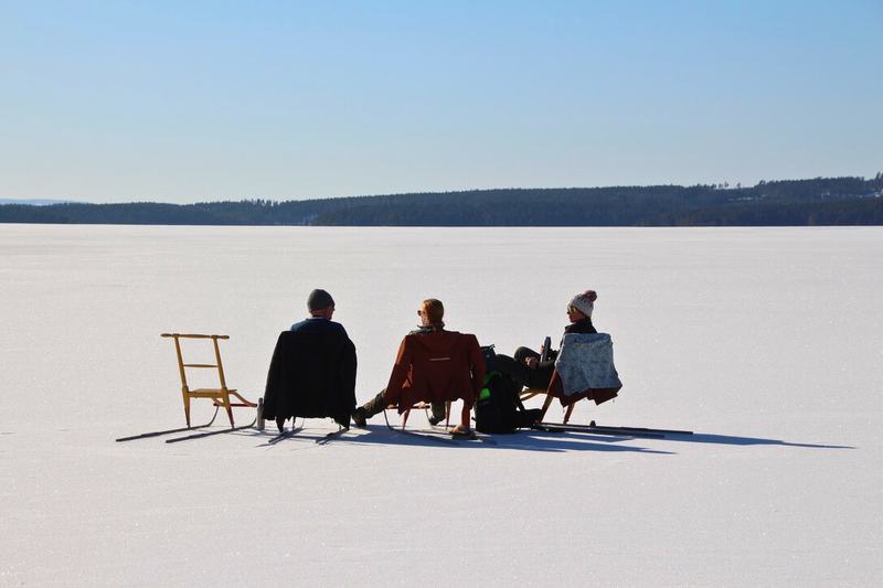 Drie mensen rusten van achteren gezien op schopsleeën op een zonnig, besneeuwd meer, met een lege houten schopslee ernaast