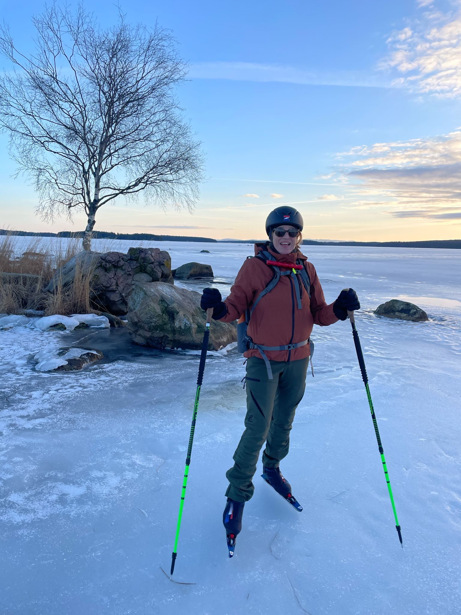 Een lachende schaatster met helm en stokken op het natuurijs bij de oever, met rotsen en een berk in de zon