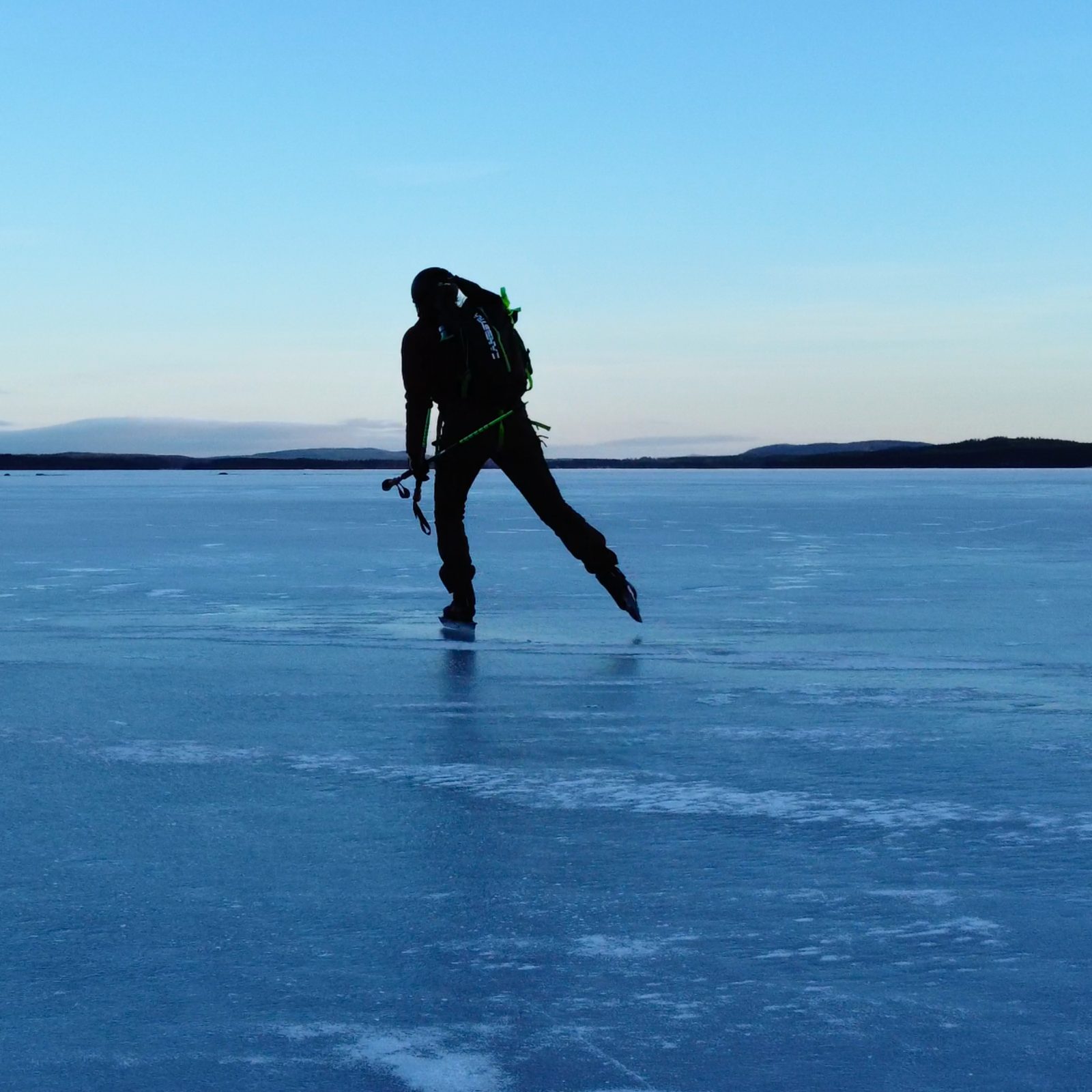 Een schaatser met rugzak en stokken op het blauwe natuurijs van een groot meer, onder een heldere lucht
