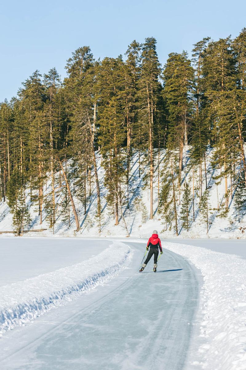 Een schaatser in een rode jas volgt een bochtige baan op het ijs onder een helling met dennenbomen