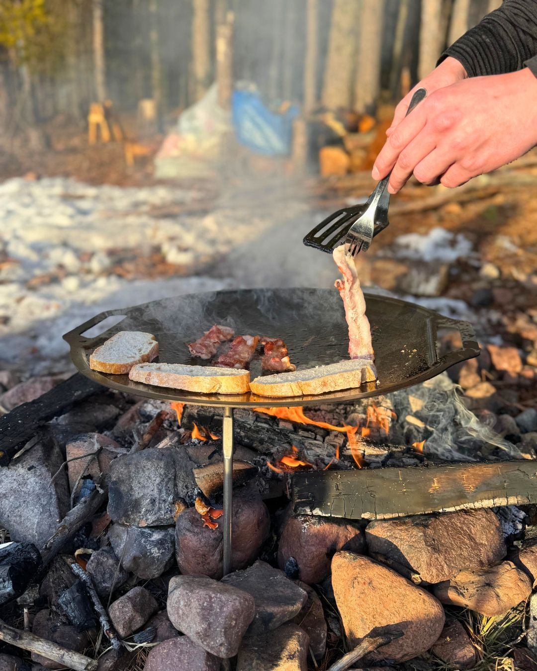 Een outdoor-lunch wordt bereid in een pan boven het kampvuur, met sneeuw en dennen op de achtergrond