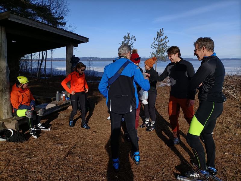 Een groep mensen in sportkleding pauzeert in de zon bij een open houten schuilhut, met een bevroren meer op de achtergrond