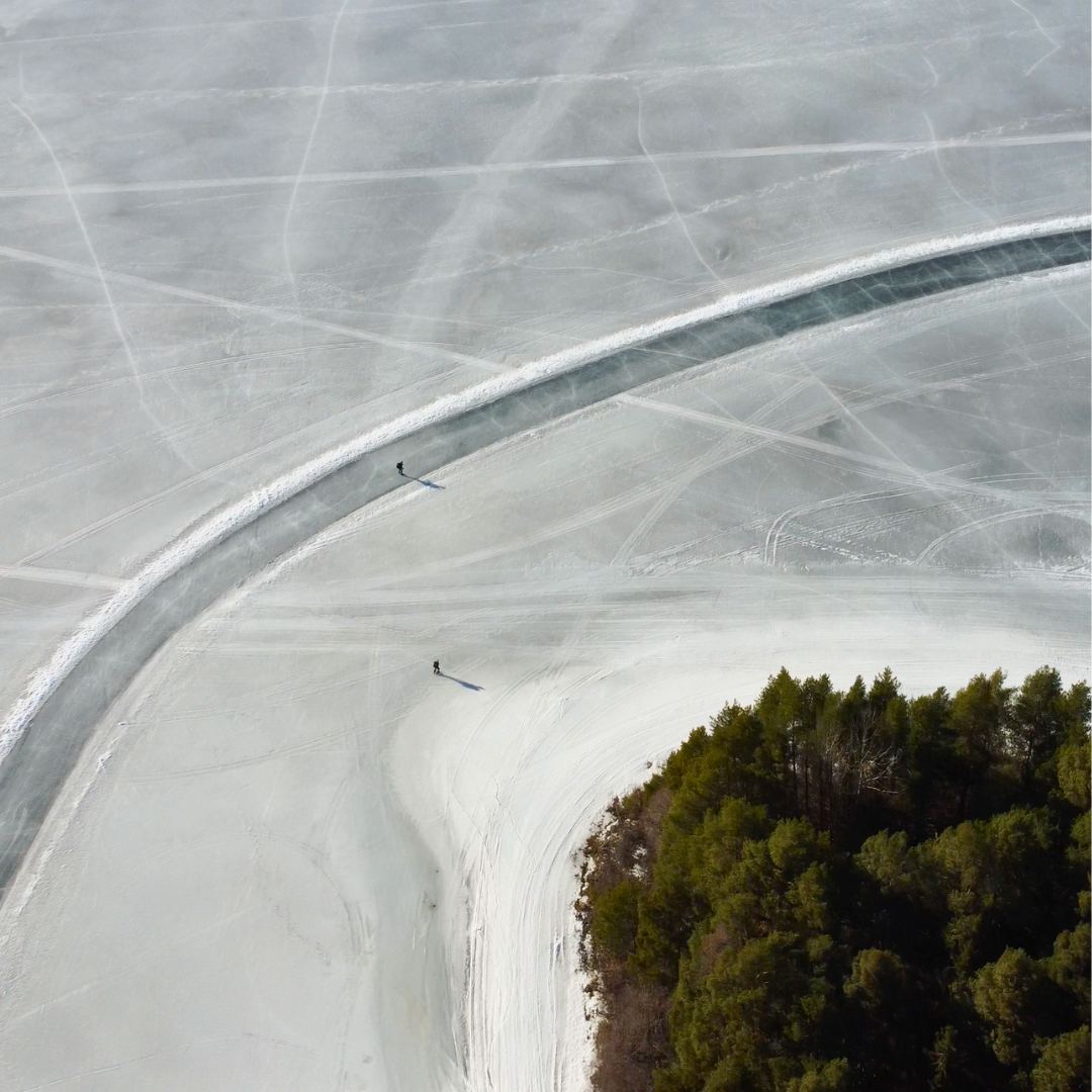 Luchtfoto van een geveegde baan die in een bocht over het bevroren meer loopt, langs een bosrijke oever, met twee mensen op het ijs