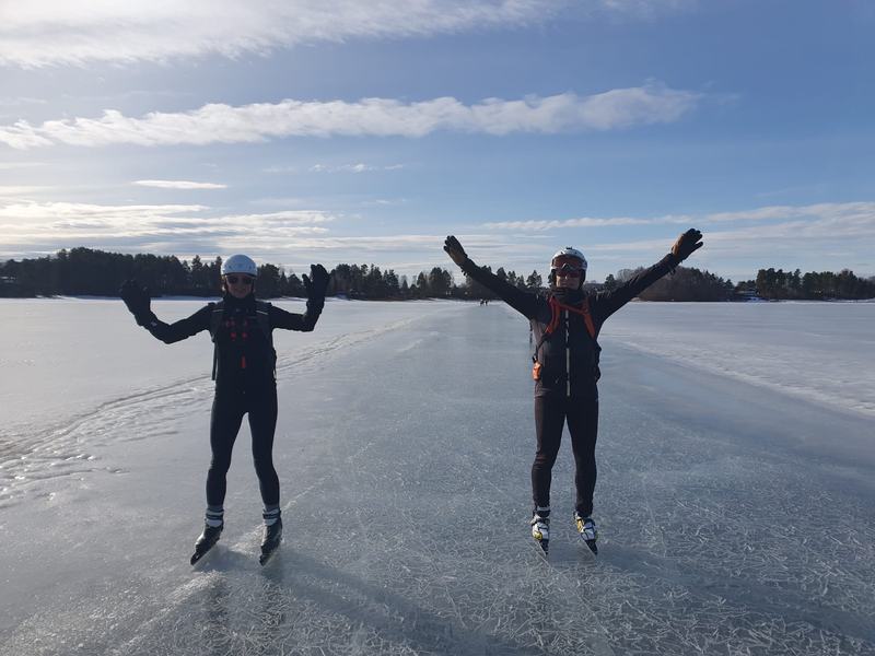 Twee schaatsers steken juichend hun armen omhoog op een bevroren meer, met tegenlicht en een bosrand op de achtergrond