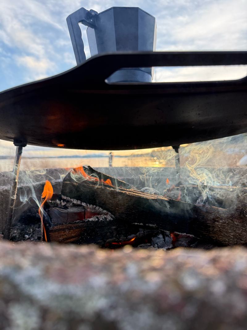 Een koffiepot boven een houtvuur in een stenen vuurplaats, met een meer en een blauwe lucht met wolken op de achtergrond