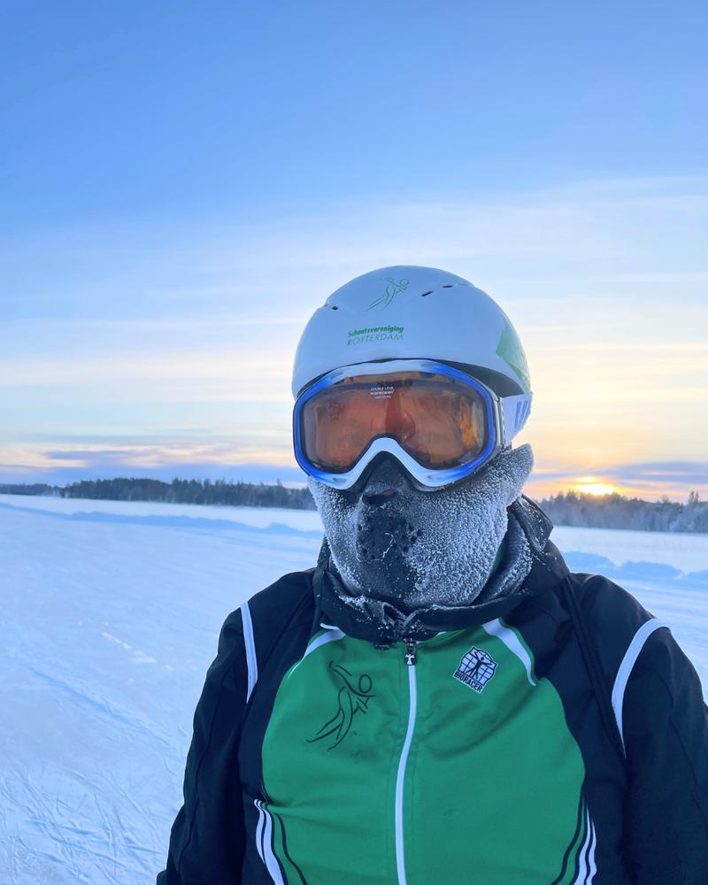 Portret van een schaatser met helm, skibril en berijpt gezichtsmasker, met een besneeuwd meer en laagstaande zon op de achtergrond