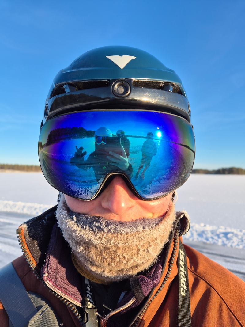 Close-up van een schaatser met helm, skibril en berijpte nekwarmer; in de spiegelende bril zijn andere schaatsers op het ijs te zien
