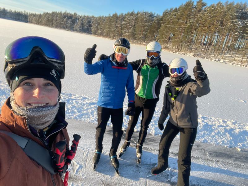 Selfie op het ijs: vooraan iemand met een skibril op het hoofd, daarachter drie schaatsers die hun duim opsteken, met een zonnig dennenbos aan de oever